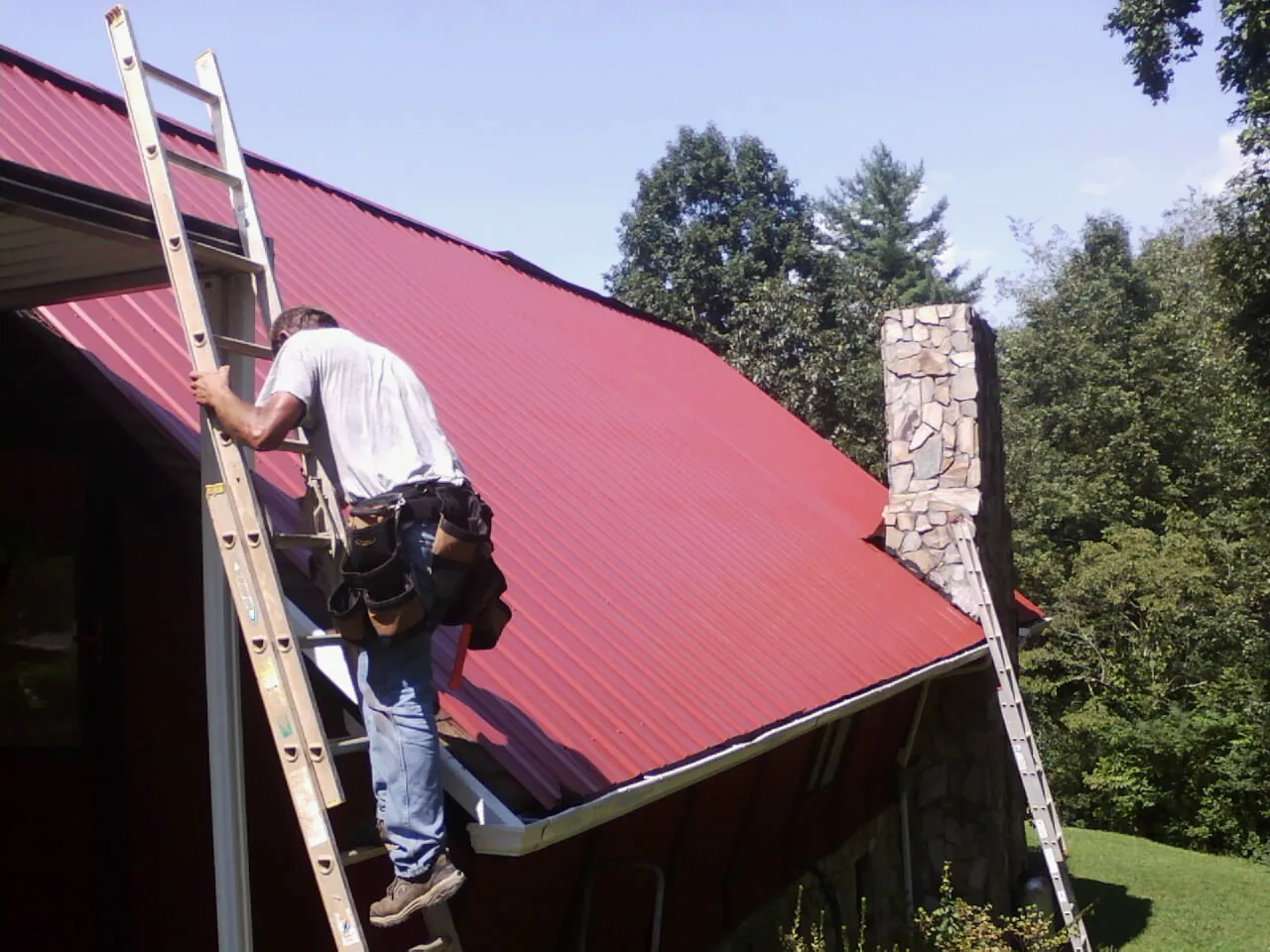 Roofer on a ladder beside a red metal roof and stone chimney — Soffit & Fascia Repair by Roofing Brothers in Macedonia